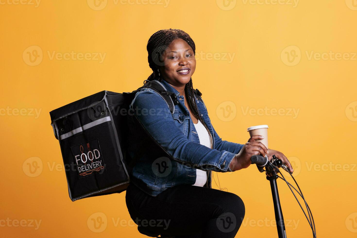 Portrait of restaurant employee courier holding cup of coffee while delivering takeaway food order to clients using bike as transportation. Pizzeria worker standing in studio with yellow background. photo