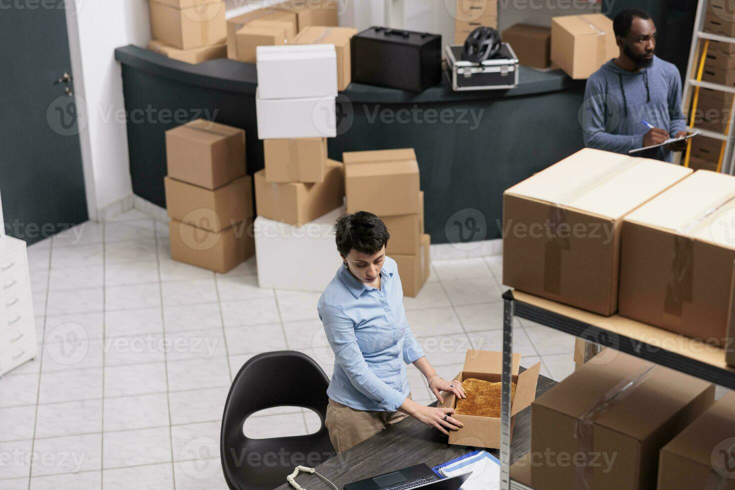 Top view of employee standing at desk in warehouse while preparing client order putting clothes in carton box after checking delivery detalies. Diverse team working in distribution center photo