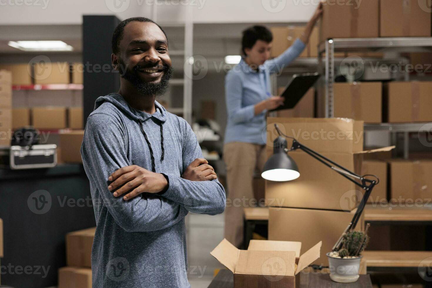 Smiling storehouse worker standing with arm crossed working at clients orders while preparing cardboard boxes in warehouse delivery department. In background woman checking packages shipping details photo