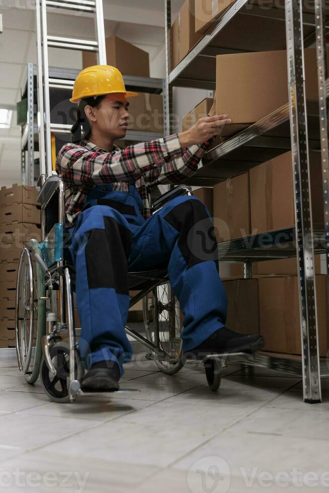 joven asiático almacén trabajador utilizando silla de ruedas mientras haciendo inventario en almacenamiento habitación. logística gerente con físico invalidez comprobación mercancías paquete o empaquetar en estante en almacén foto