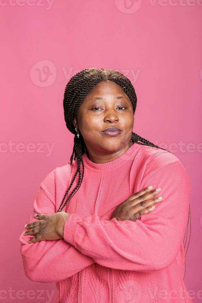 Portrait of woman smiling with confidence while posing in studio having joyful expression enjoying shoot time. African american model with stylish hairstyle and pink sweather looking at camera photo