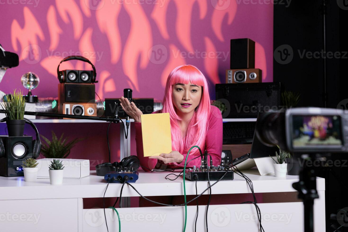 Vlogger with pink hair holding notebook advertising school supplies in front of camera while filming product review using vlogging equipment. Influencer live streaming in broadcast studio photo