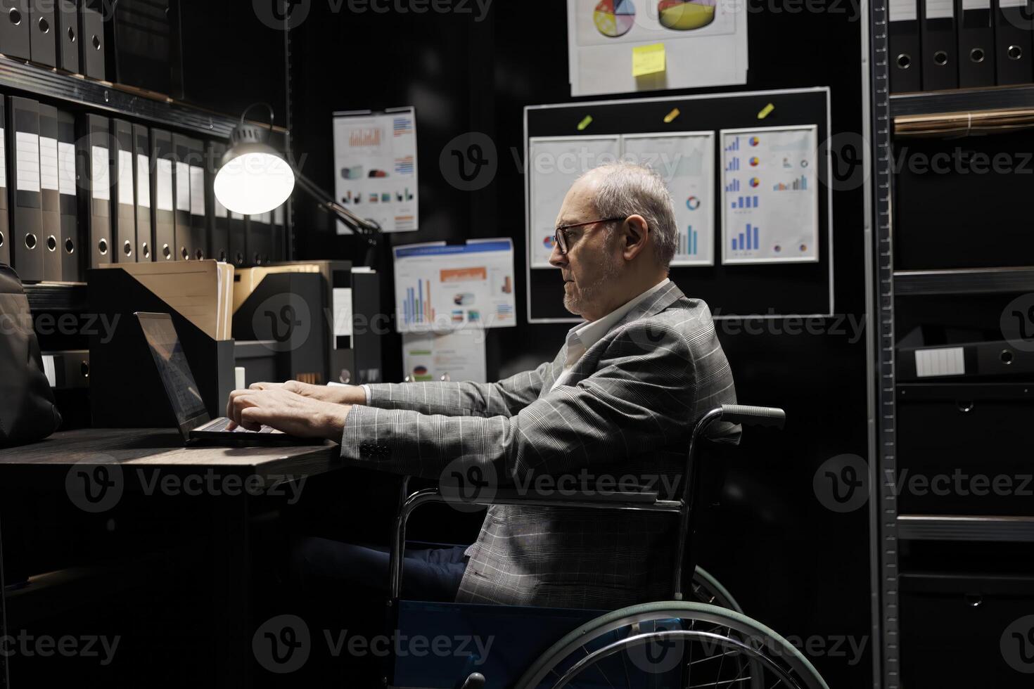 Senior businessman employee with disability in wheelchair inserting accountancy budget plan data on laptop. Bureaucratic executive in storage room office filled with chart reports and document folders photo