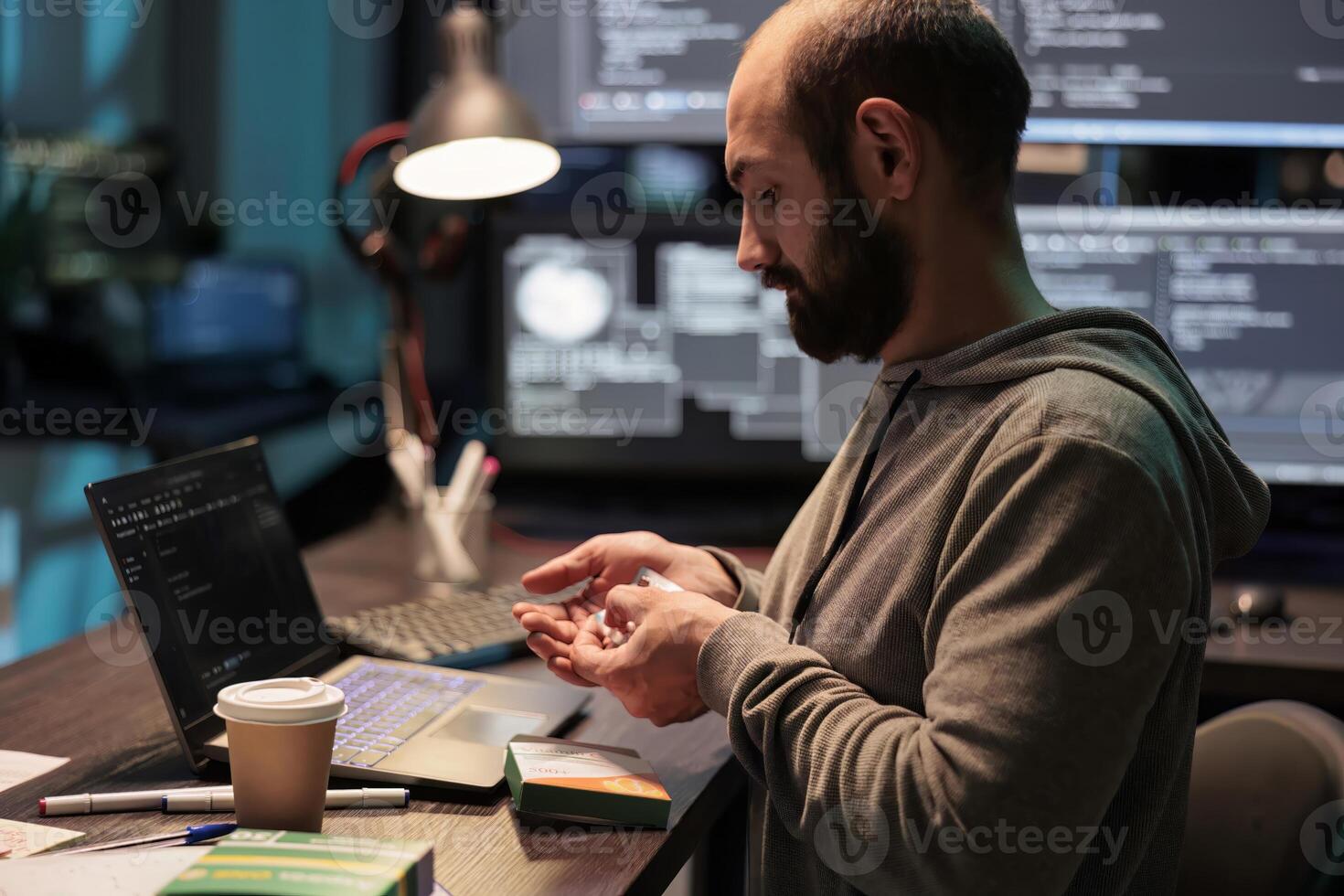 IT engineer taking vitamins and working after hours, using medicine and supplements to stop being tired or stressed. Taking medicaments from foil and box, programming source code. photo