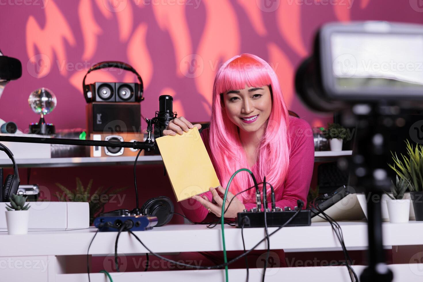 Influencer with pink hair advertising notebook in front of camera recording vlog while presenting school supplies review using professional vlogging equipment. Asian creator filming podcast photo