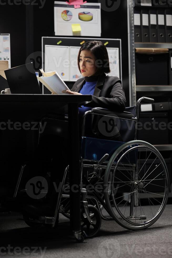 Paralyzed professional asian accountant in wheelchair checking administrative paperwork in file room. Businesswoman in bureaucratic file cabinet office filled with invoice documents and flowcharts photo