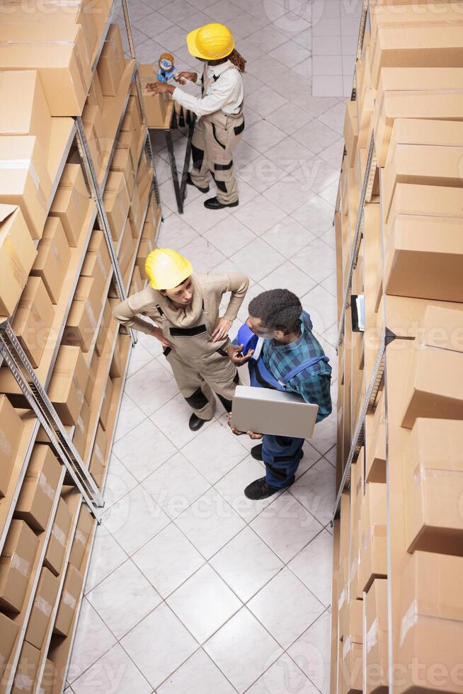 Warehouse supervisors analyzing stock management system on laptop and packing parcels with adhesive tape. Shipping company employees preparing cardboard boxes for sending and chatting top view photo