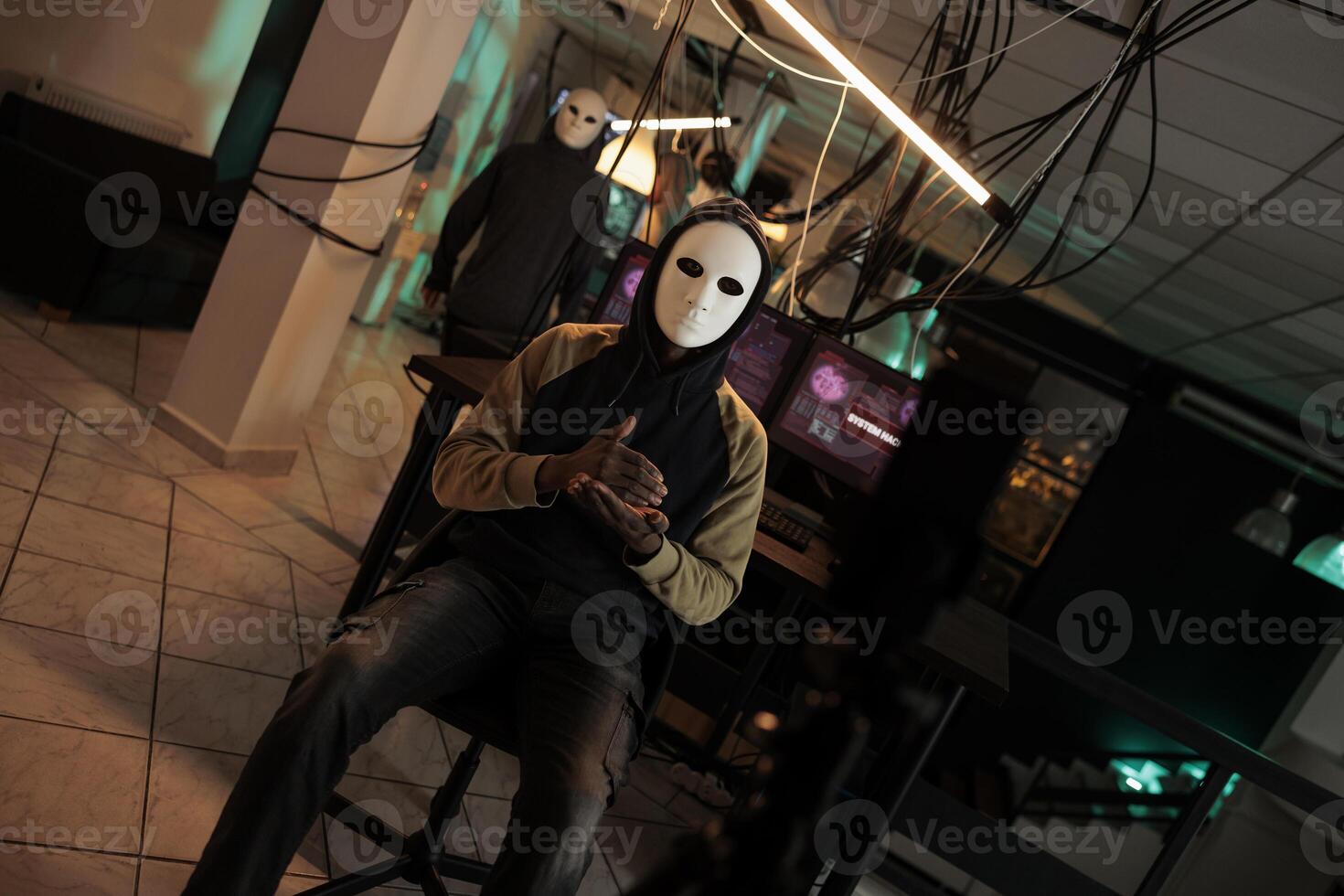 Criminal with anonymous mask threatening to expose government data on live broadcast, asking for ransom at night. Young thief hacking computer system and breaking into security server. photo