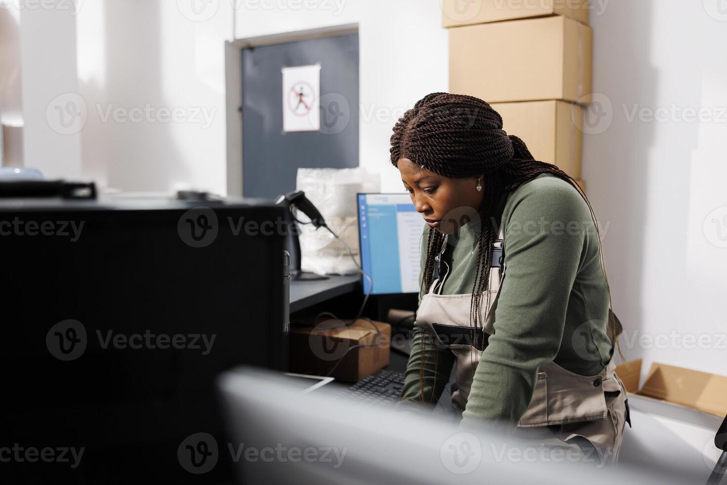 African american supervisor analyzing warehouse logistics on laptop computer, working at merchandise inventory in storage room. Storehouse employee preparing customers orders for shipping photo