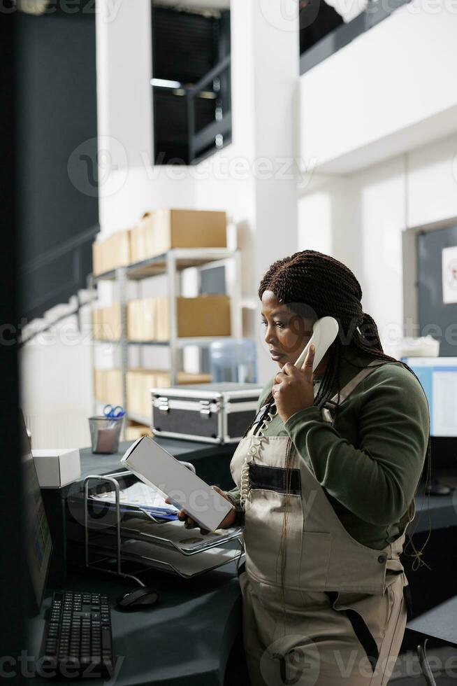 African american worker looking at white cardboard box while talking with remote client, discussing online order using landline phone. Stockroom supervisor working at goods inventory photo