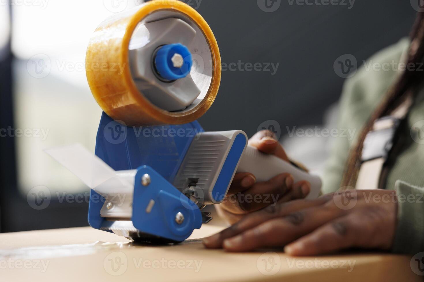 African american manager preparing customers orders, using adhesive tape to pack products in cardboard boxes in warehouse. Storage room worker wearing industrial overall during inventory. Close up photo