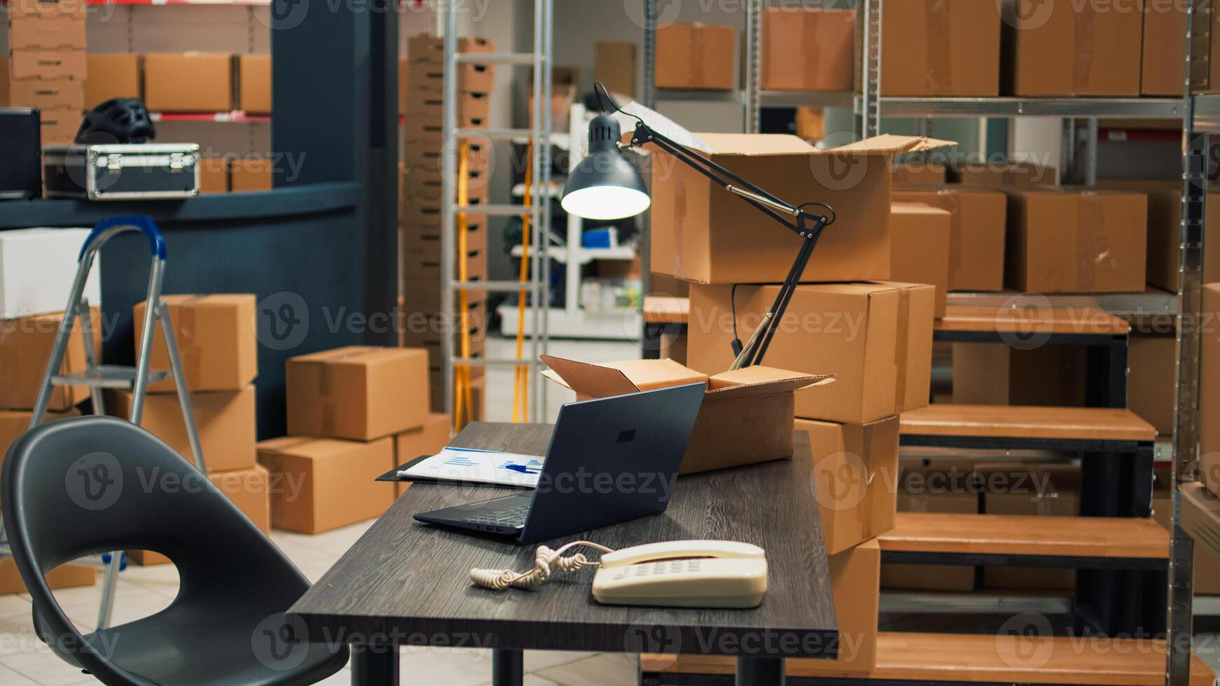 Empty desk room in storage warehouse area filled with carton boxes, office with laptop and inventory or logistics notes. Storespace with stacks of merchandise in cardboard packages. photo