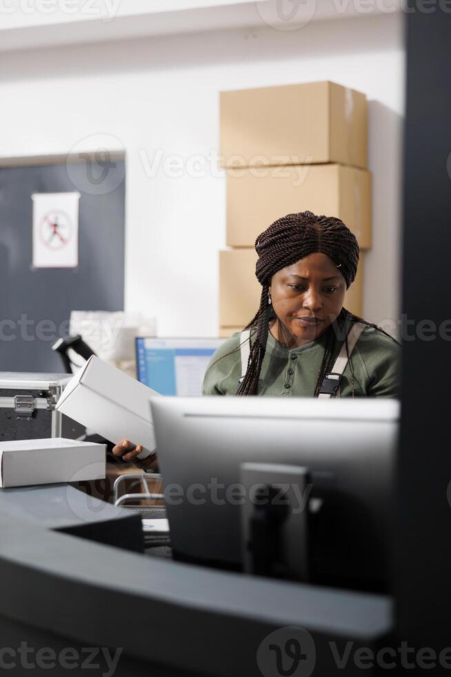 African american supervisor holding white carton box, preparing clients orders for shipping in warehouse. Small business worker wearing industrial overall working at inventory in storage room photo
