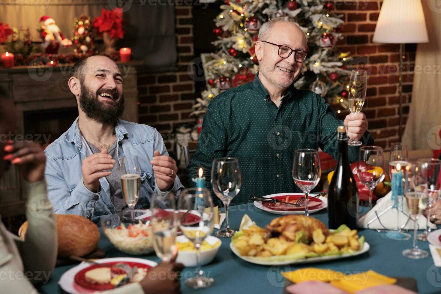 mayor hombre haciendo brindis con elevado vaso de vino a Navidad víspera cena a celebrar diciembre fiesta con amigos y familia. abuelo haciendo habla alrededor el mesa, espumoso alcohol. foto