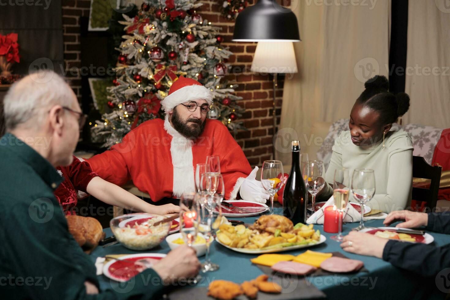 Jolly santa claus making people laugh at festive dinner, celebrating christmas eve holiday together at home. Senior couple, diverse friends and family gathering near santa to have fun. photo