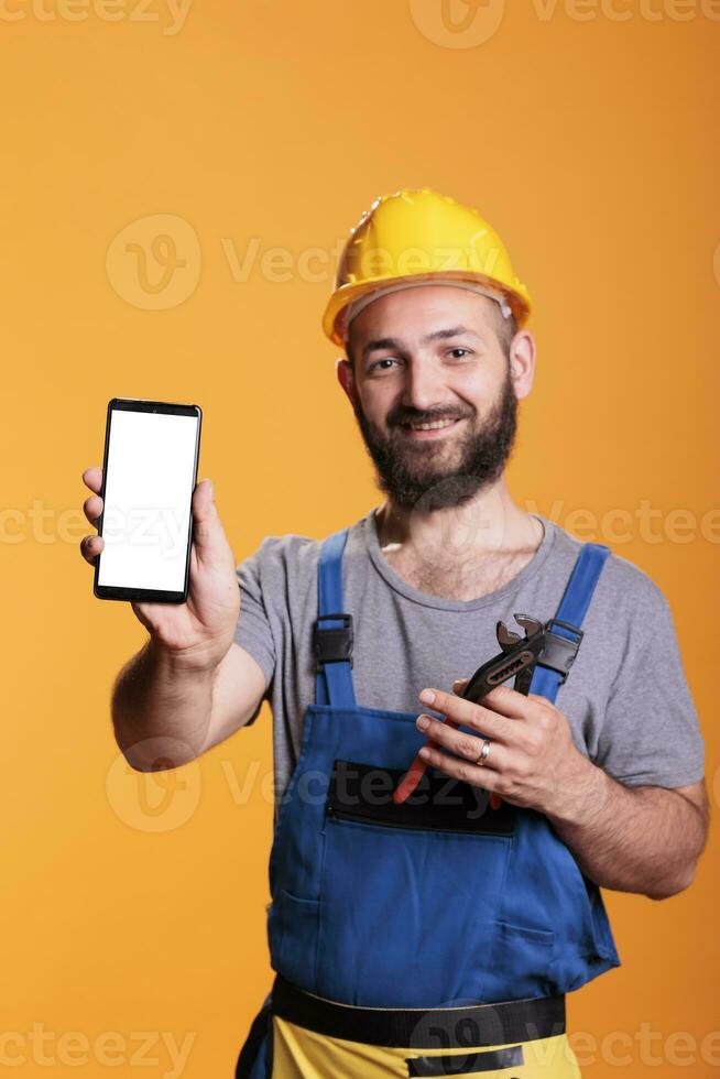 Construction worker holding cell phone with empty screen for advertising in front of camera. Professional builder with hard hat against yellow background in studio shot. photo