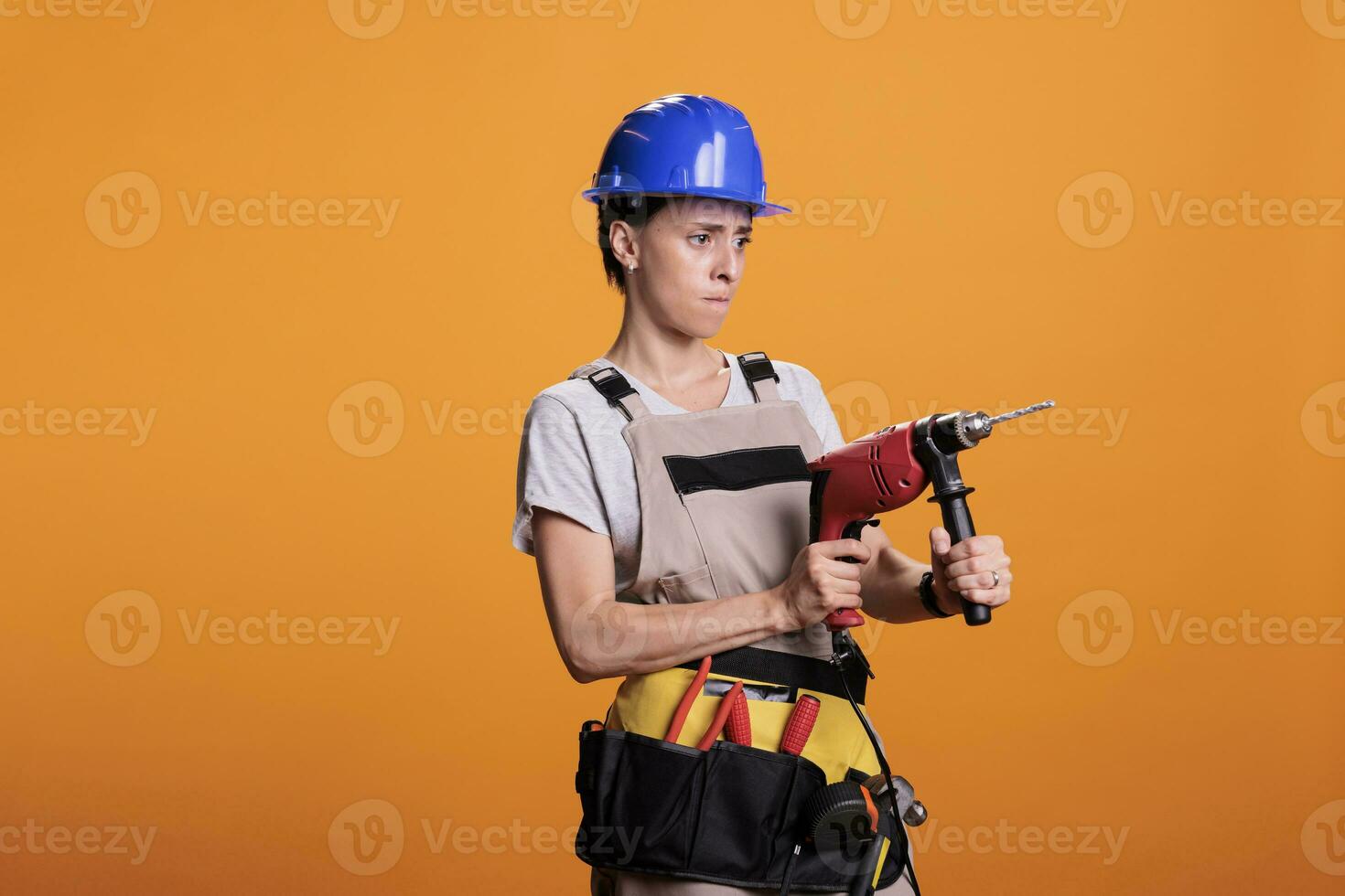 Professional female builder holding drilling gun in new interior renovation project, using power drill nail gun. Woman renovator wearing uniform in studio shot over yellow background. photo