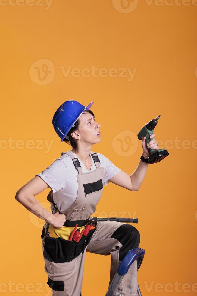 Woman working as builder on ladder drilling wall with cordless power drill gun. Professional female construction worker in studio over yellow background, building refurbishment. photo
