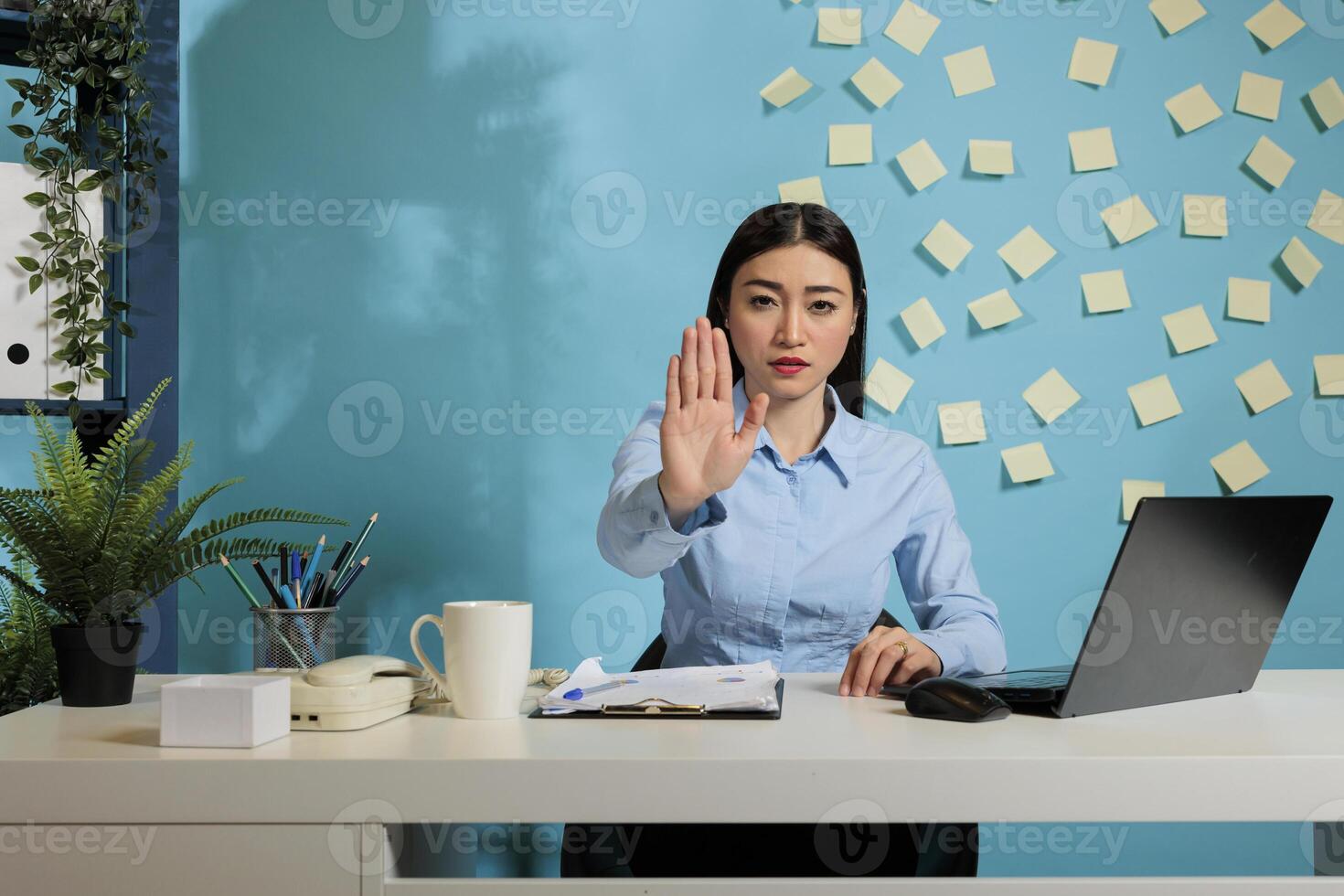 Woman seated at office desk showing rejection with the palm of her hand, expressing dissatisfaction with working conditions. Employee refusing to receive more workload without rest. photo