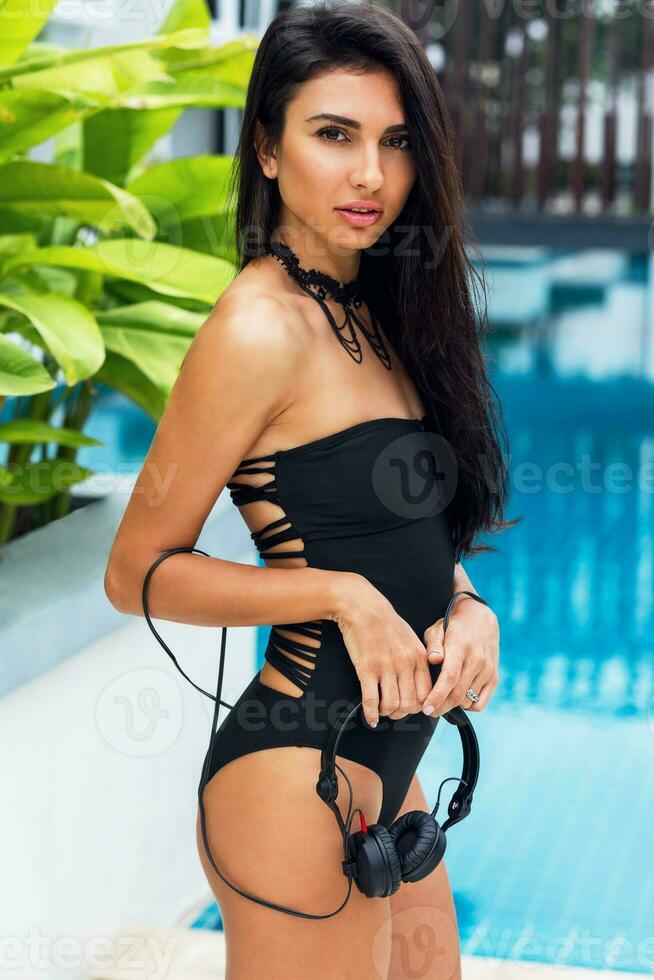 Sensual woman with perfect slim tan body in trendy necklace , black swimwear and transparent beach cover up , trendy yellow sunglasses  enjoying  holidays  near amazing pool in tropical resort. photo