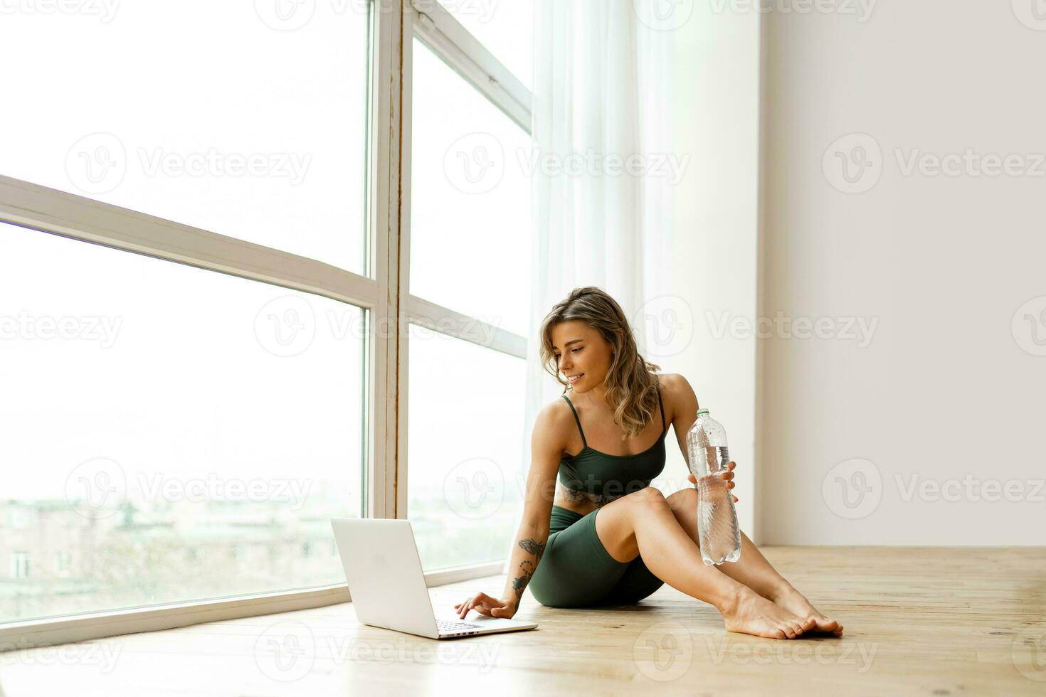 Pretty  woman in sportswear is sitting on the floor with  bottle of water and is using a laptop  in  modern fitness  studio . Sport and recreation concept. photo