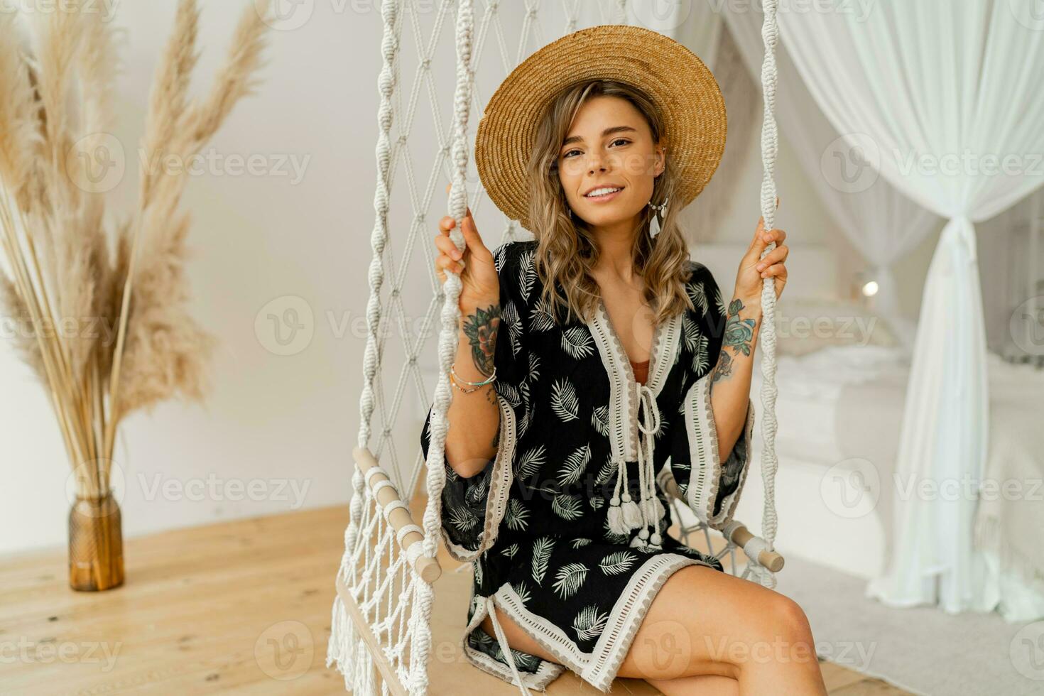 Smilling young woman in boho dress posing in stylish bedroom.  Sraw hat, boho feather drop earrings . Lady sitting on hanging swing. photo