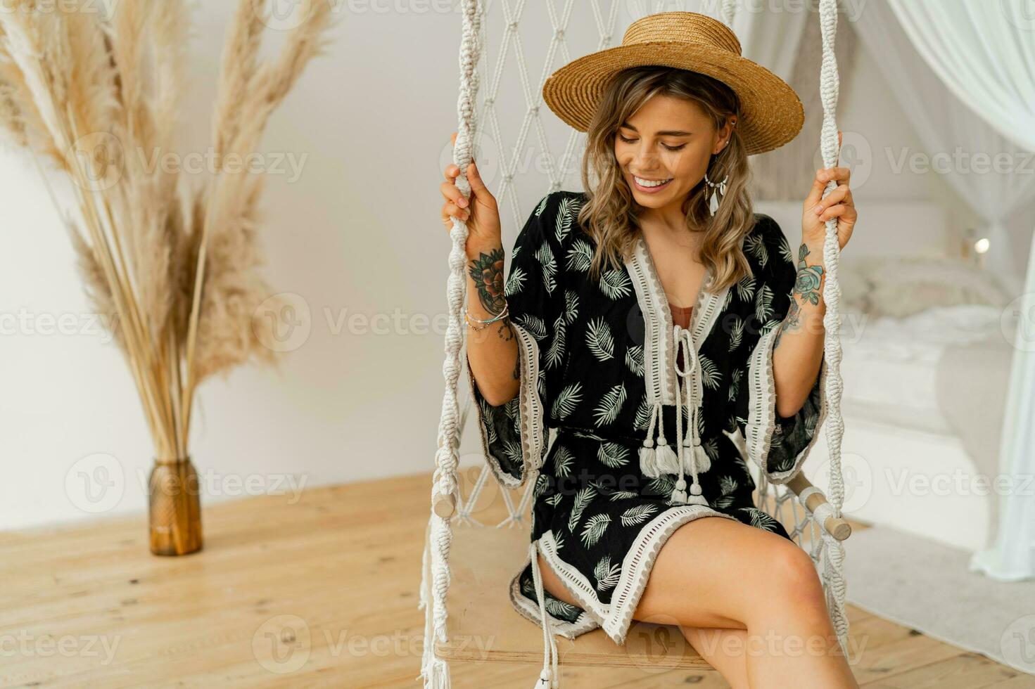 smilling joven mujer en boho vestir posando en elegante dormitorio. crudo sombrero, boho pluma soltar pendientes . dama sentado en colgando balancearse. foto