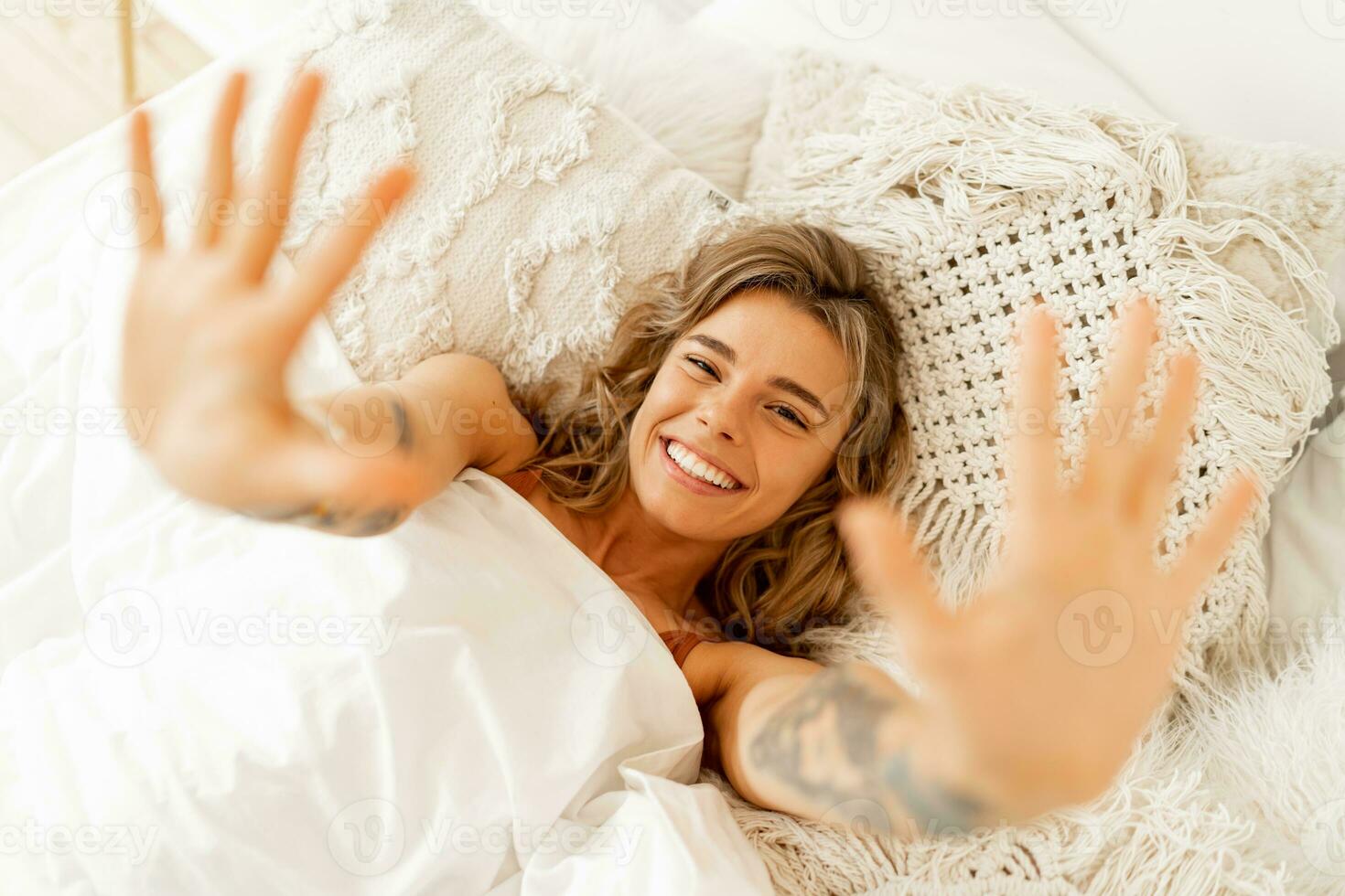 Top view portrait of happy cheerful  woman lying in white cozy bed wake up in the morning, pulls  hands forward. Warm colors, boho interior. photo