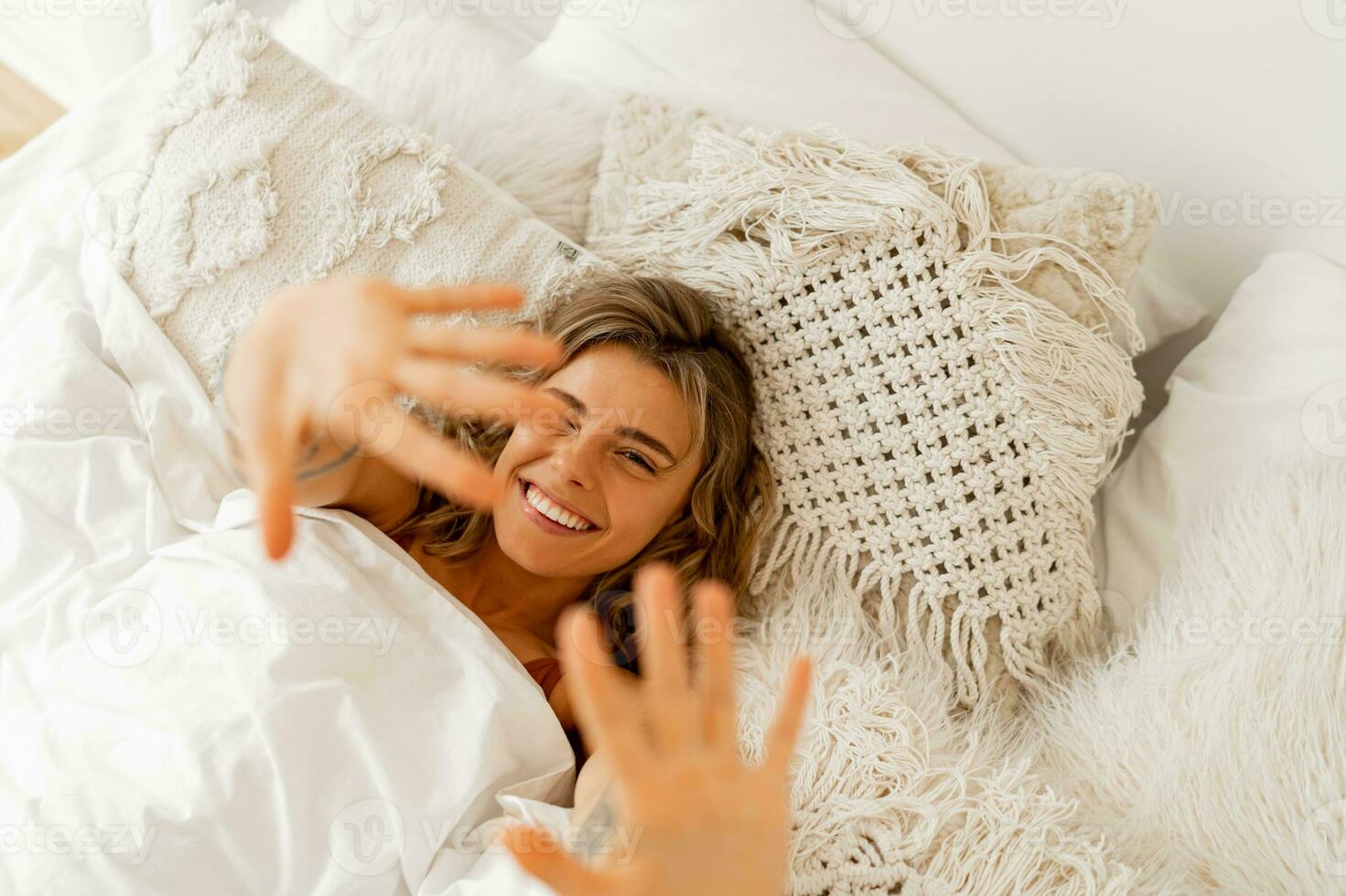 Top view portrait of happy cheerful  woman lying in white cozy bed wake up in the morning, pulls  hands forward. Warm colors, boho interior. photo