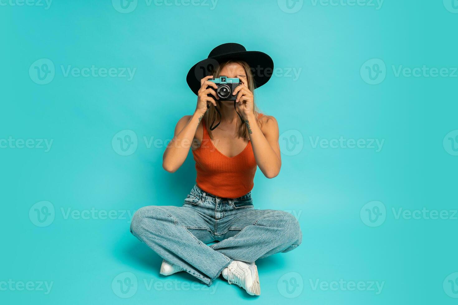 Lovely blond woman in  stylish summer outfit holding retro camera , sitting on floor in studio on blue background. Vacation mood. photo