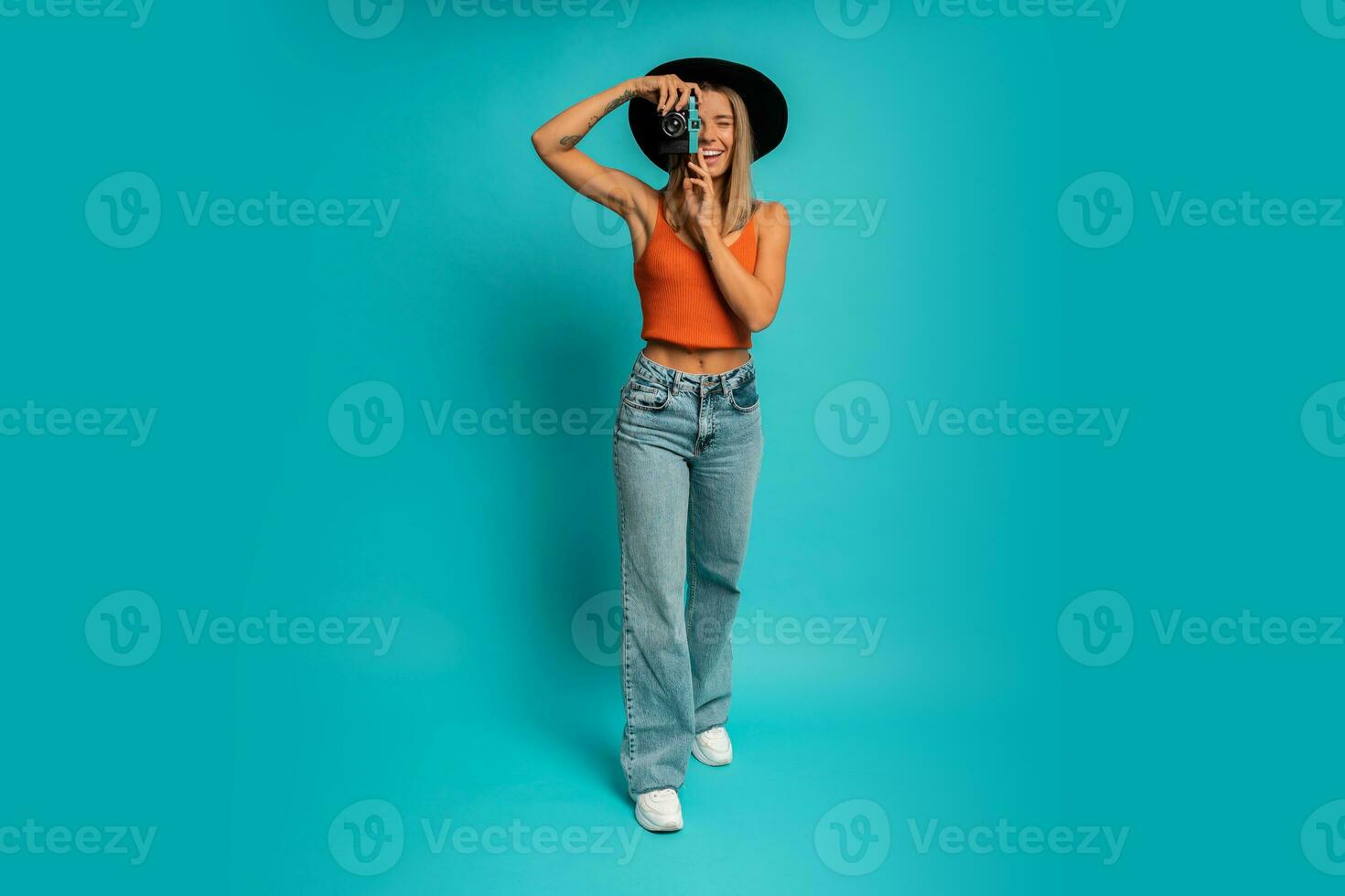 Pretty laughing woman in black hat and orange top  holding vintage camera and posing over turquoise background in studio.  Full lenght. photo