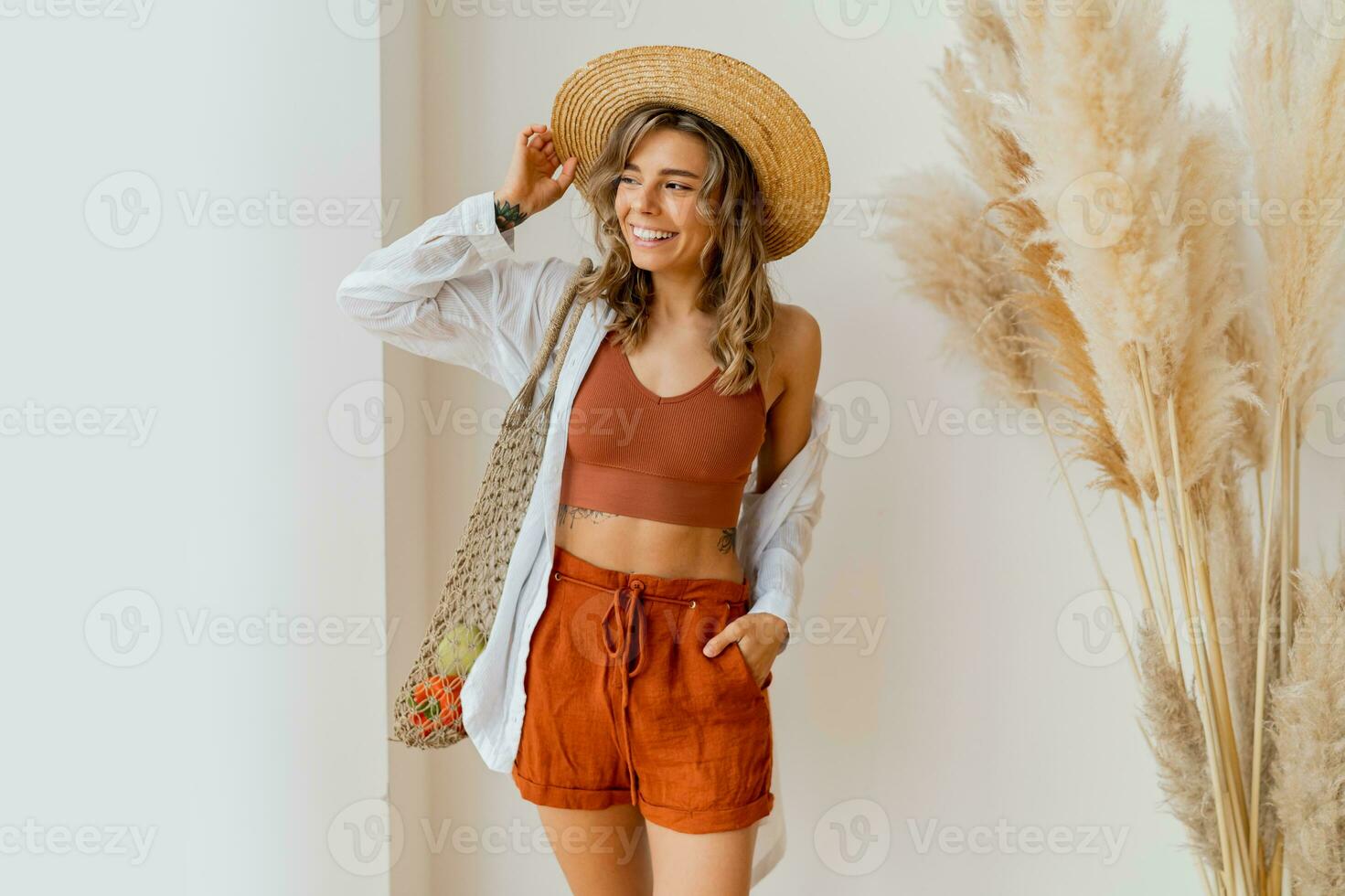 Pretty blond woman in straw hat  holding eco friendly mesh shopper with fresh  vegetables  in studio with boho interior. photo