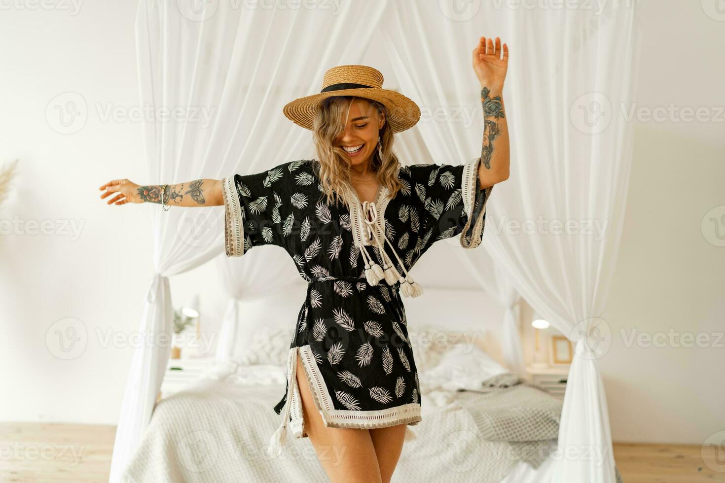 Happy woman dancing and having fun in cozy  bedroon boho style.  Wearing straw hat and  summer dress. photo