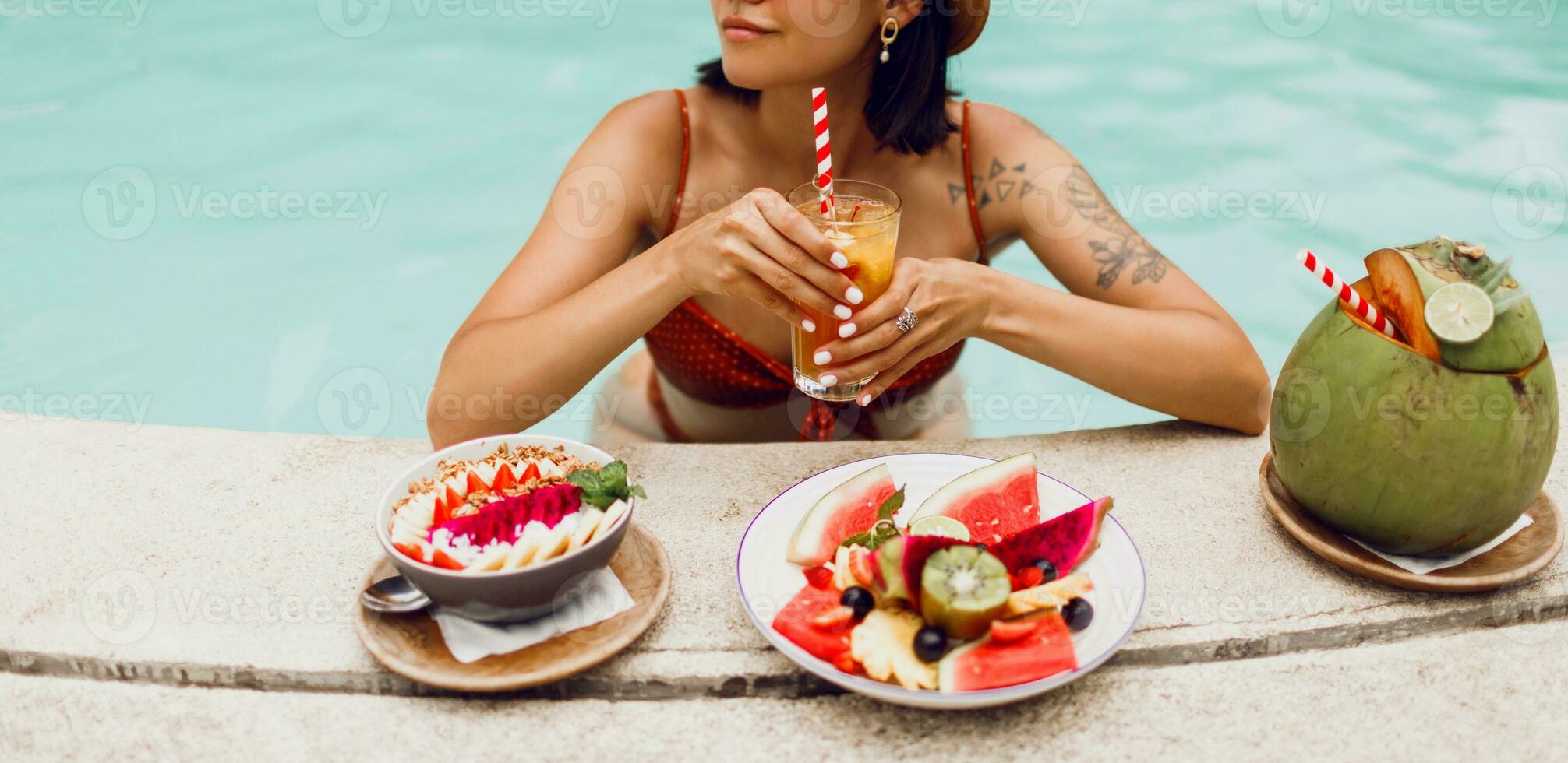 Confident  brunette tan woman in red cat eyes sunglasses   relaxing in   pool  with plate of exotic fruits  during tropical vacation.  Stylish tattoo. photo