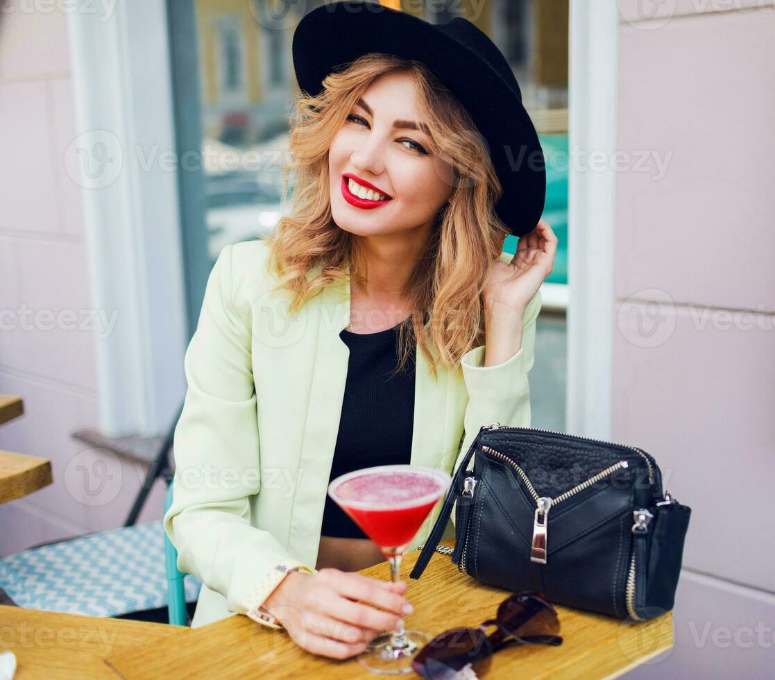 Stylish woman in casual summer outfit in black hat sitting in modern cafe and drinking Cocktail. photo