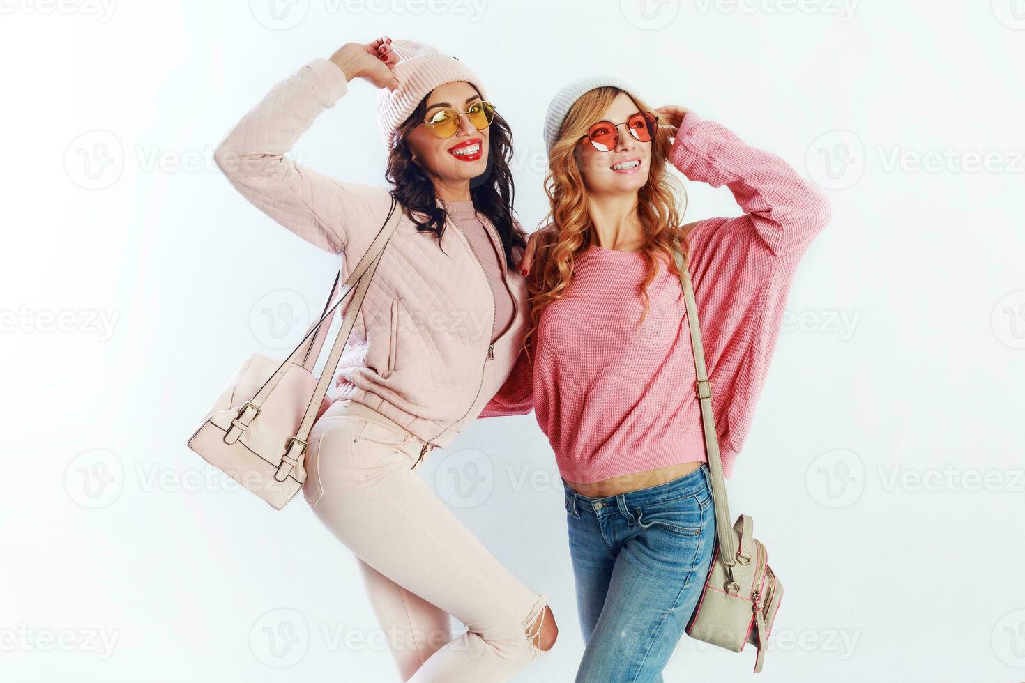 Indoor studio image of two girls, happy friends in stylish pink clothes and hat spelling funny  the together. White background. Trendy hat and glasses.Showing peace. photo