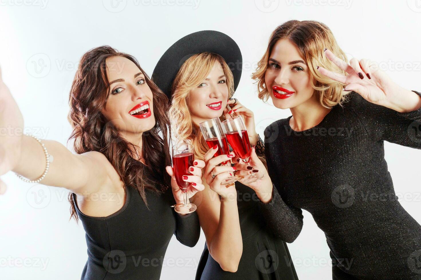 Close up self portrait of Three  pretty women  celebrate hen-party and drinking cocktails. Best friends wearing black evening dress and heels.  Bright make up, red lips. Inside. photo