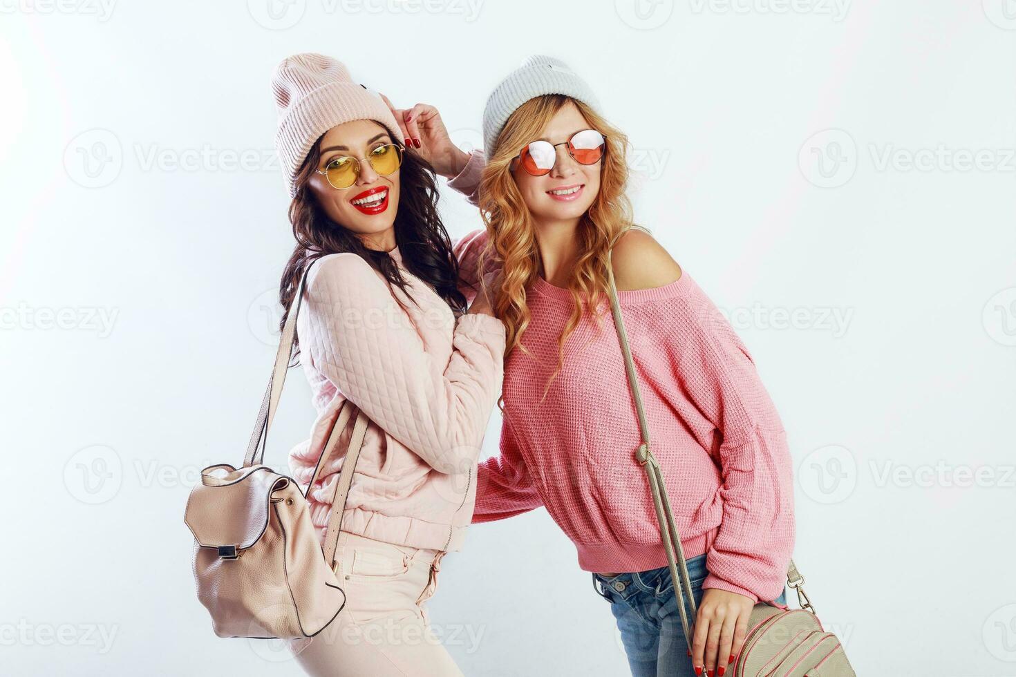 Indoor studio image of two girls, happy friends in stylish pink clothes and hat spelling funny  the together. White background. Trendy hat and glasses.Showing peace. photo