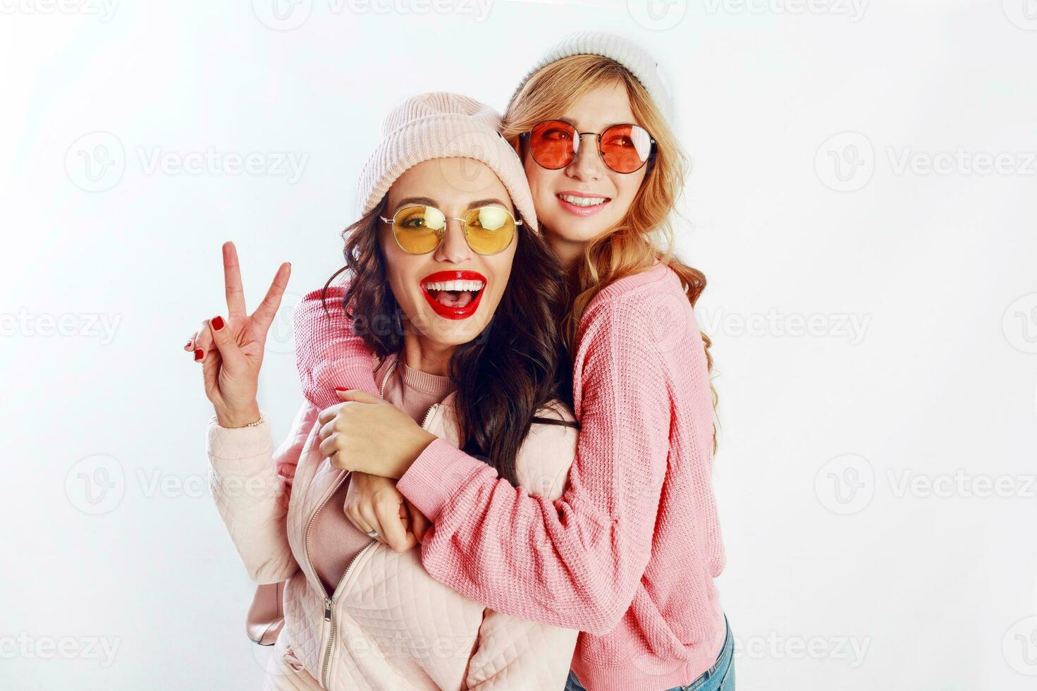 Indoor studio image of two girls, happy friends in stylish pink clothes and hat spelling funny  the together. White background. Trendy hat and glasses.Showing peace. photo