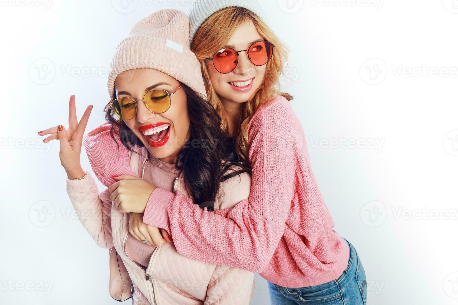 Two cheerful young friends in perfect mood having time together on white isolate background. Positive girls wearing pink  cozy sweaters and pullover, trendy hat , cute glasses. photo