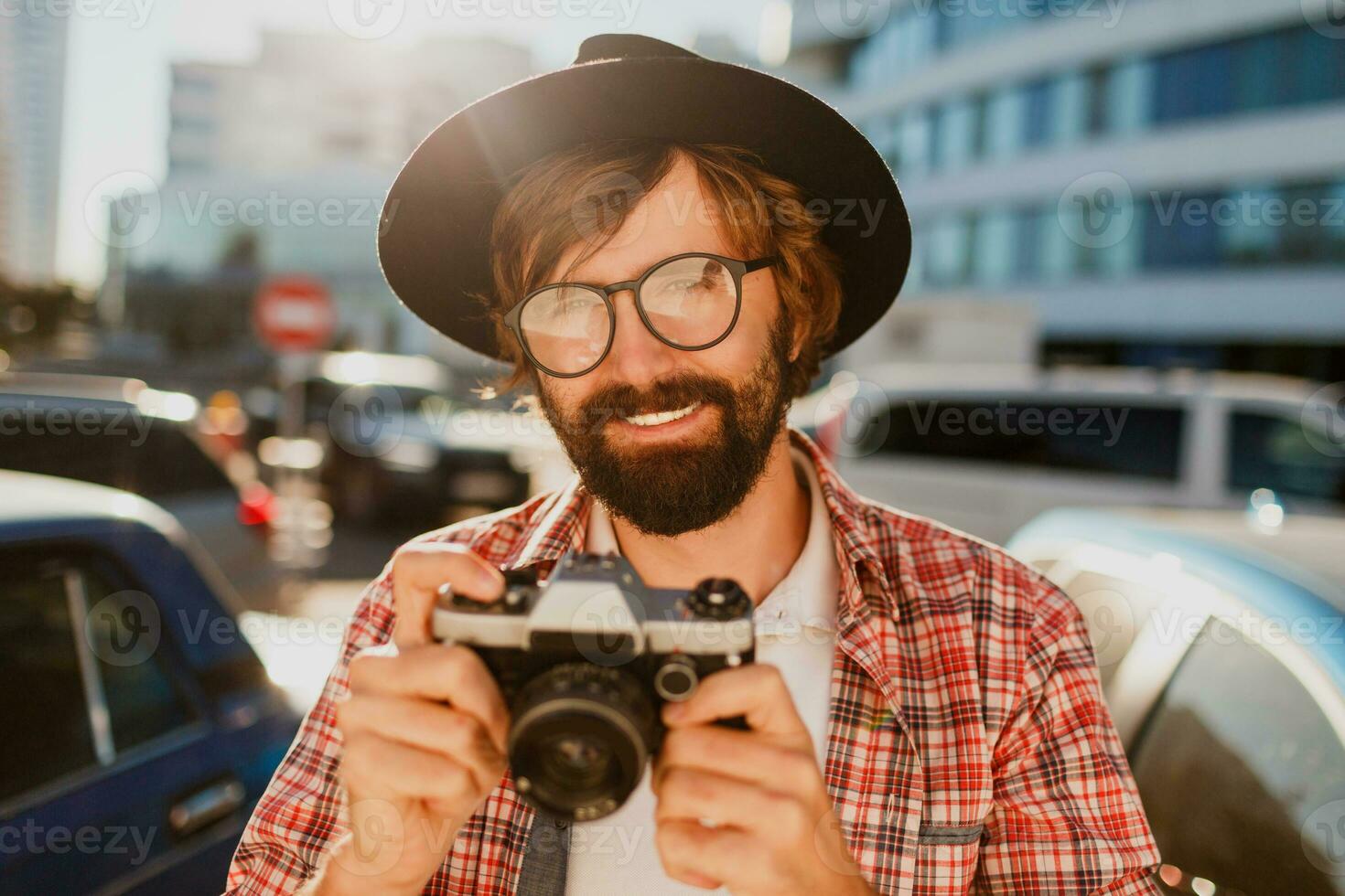 Close up  portrait of smiling  Hipster   beard man using retro film camera, making  photos while traveling. Urban background. Stylish spring outfit. Wearing black hat, checkered shirt .