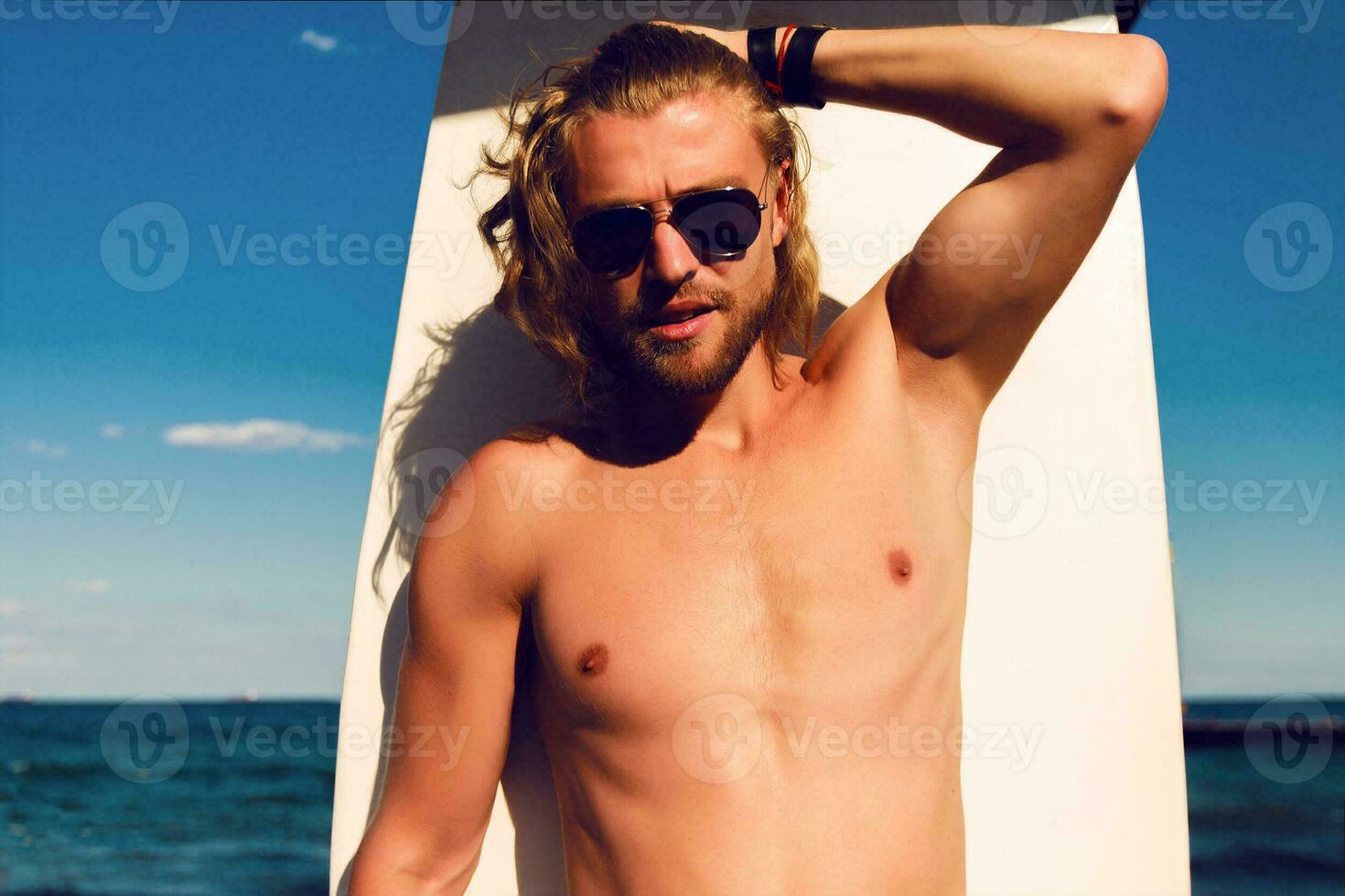 Young attractive surfer sports man  with long hair wearing jeans shorts   and looking at the horizon while carrying his surfing board during a sunny day against the intense blue sky. photo