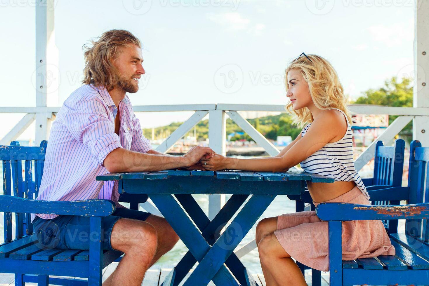 Attractive  guy and beautiful blond girl having a date  sitting on blue chairs in retro beach cafe and enjoying summertime . photo