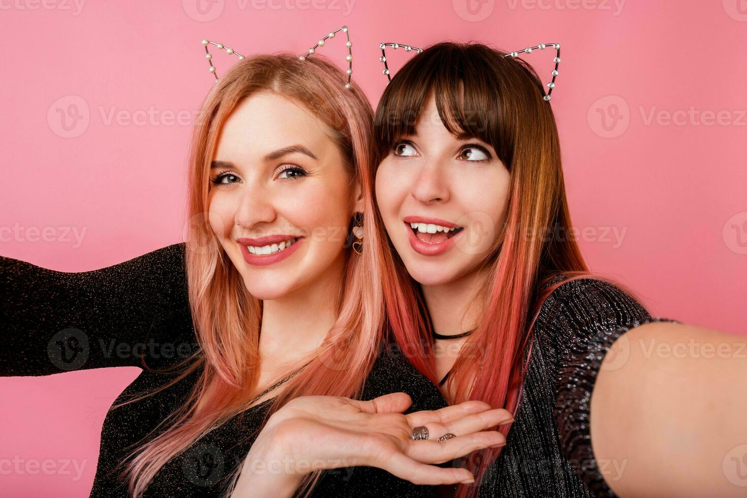 Two beautiful women in masquerade cat ears  making self portrait on pink background. Close up portrait  of embracing girls with candid smile. Party mood. photo