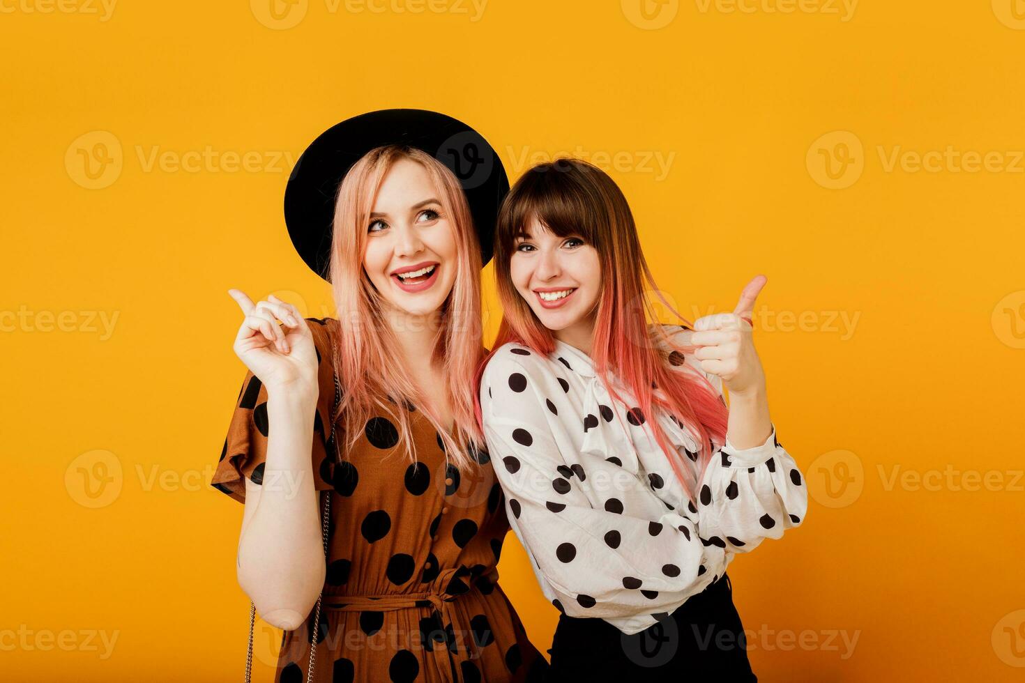 Indoor positive portrait of two gorgeous girls posing over yellow background. Vintage outfit, black hat, candid smile. Party mood. photo