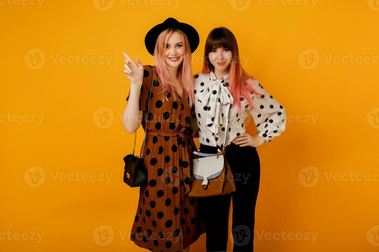 Two friends, pretty girls walking and having fun on yellow background in studio. Happy woman with pink hairs  with her blond friend  having great time. photo