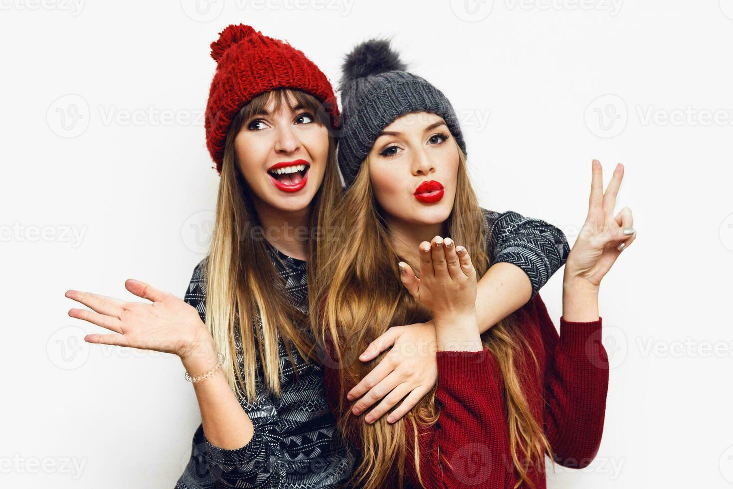 Indoor lifestyle  portrait of two pretty happy  girls, best friends in cute knitted hats and cozy sweaters having fun, send kiss to camera, surprised faces on white wall. Flash. Isolate. photo