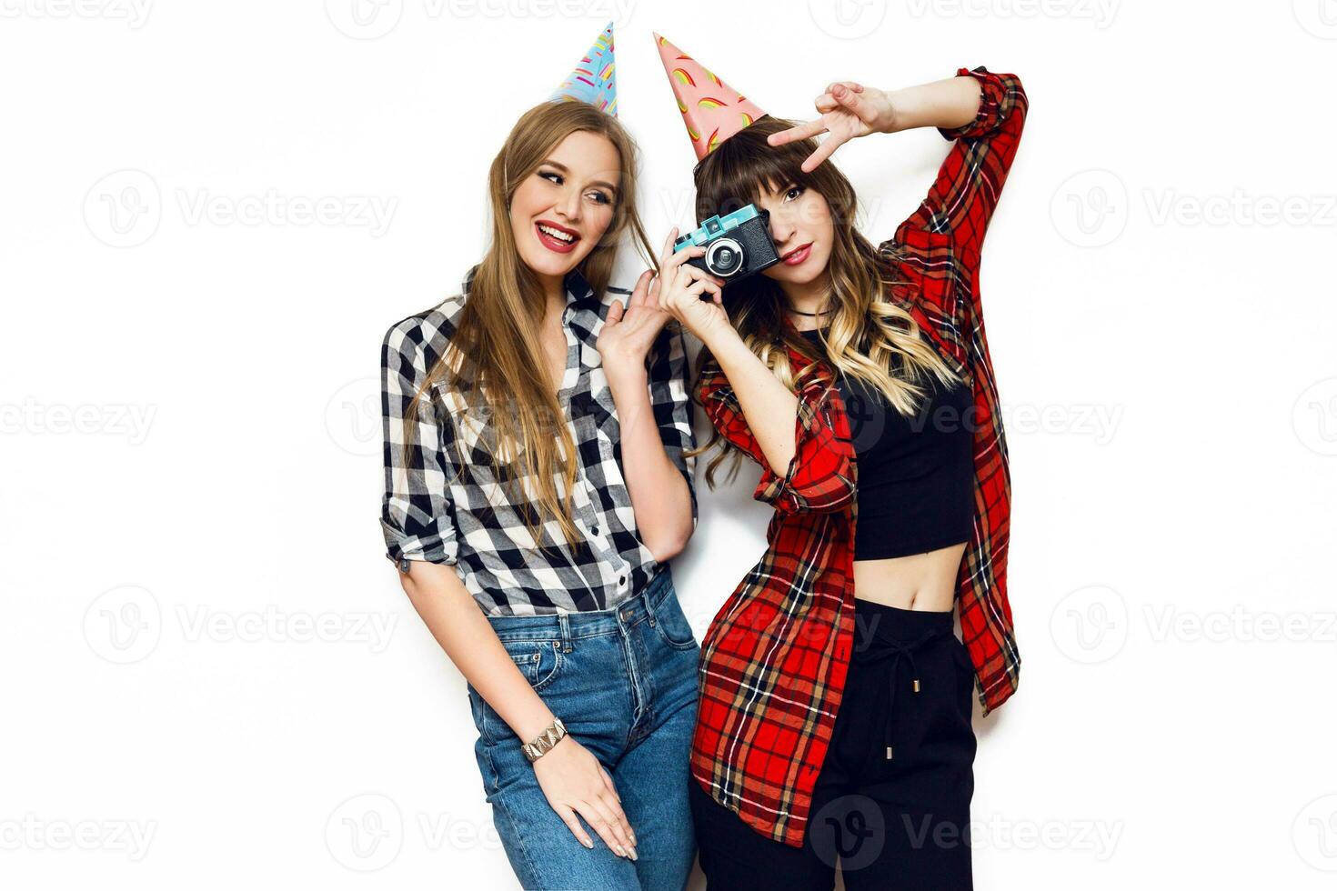 Two  cheerful friends, cute girls having fun together, posing on white background. Wearing stylish party hat , making  photos, send kiss, laughing , emotions,  party time, celebrating  birthday. photo