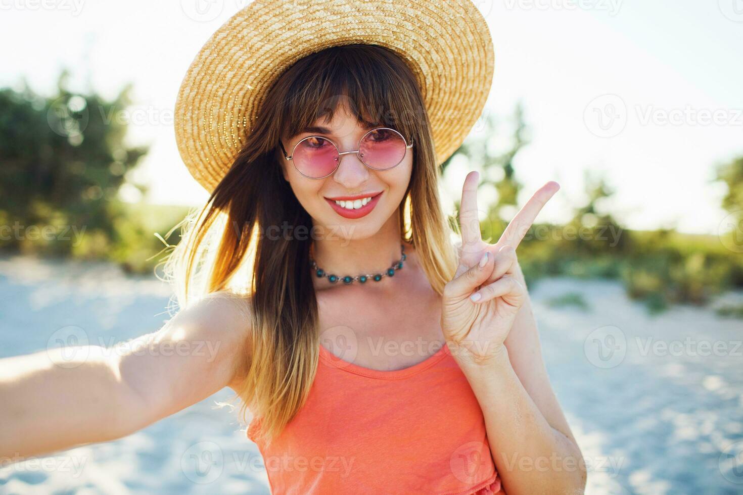 elegante bonito joven mujer en linda rosado Gafas de sol , Paja sombrero y de moda verano atuendo haciendo selfie en cámara , demostración señales , reír. foto