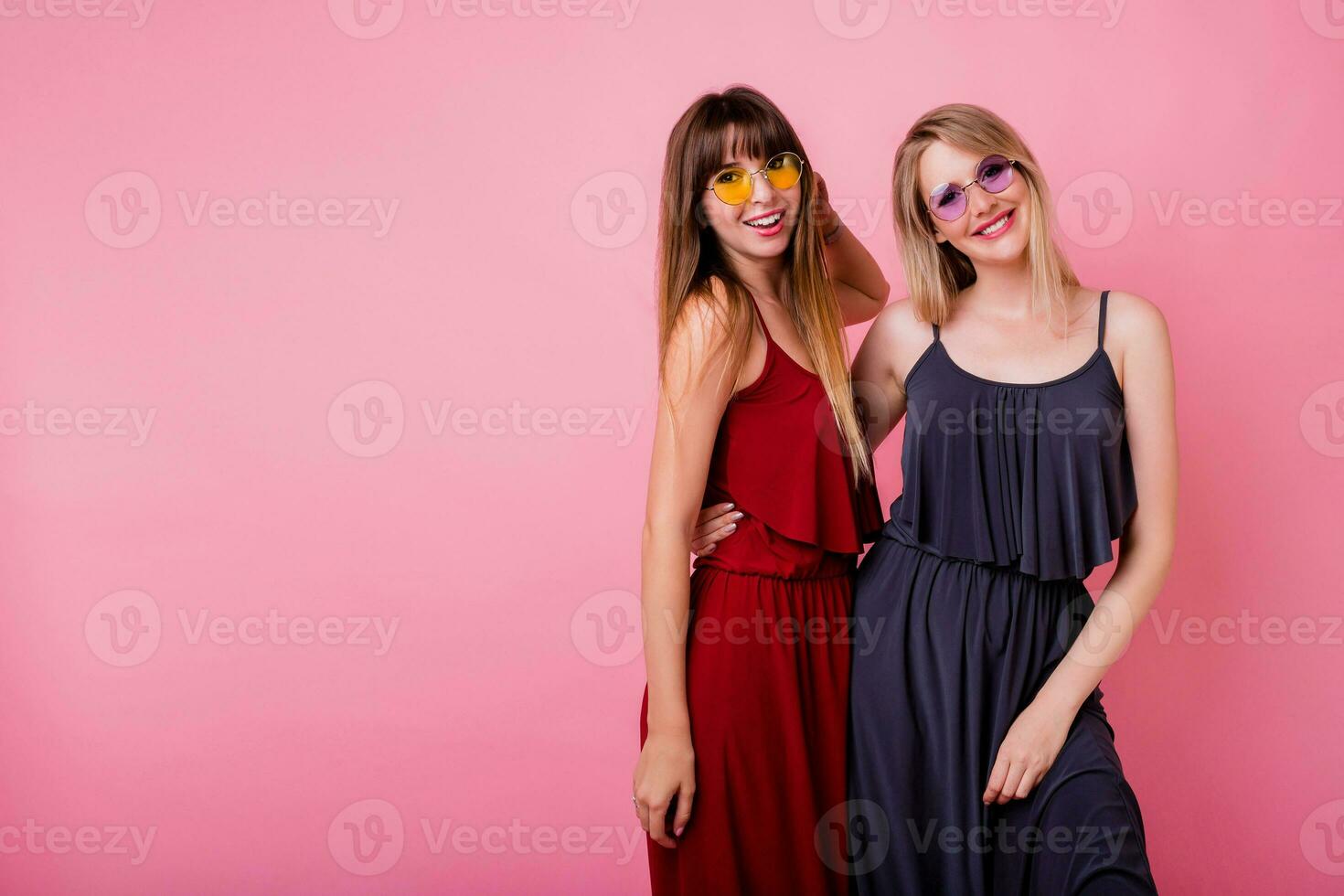 Couple of smiling women posing on pink background. Two smiling friends  hugging and looking on camera. Stylish similar dress. photo